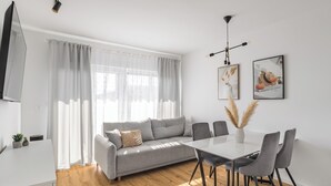 A modern living room with a grey sofa, a dining table with chairs, and a TV on the left. - Did' (Split)
