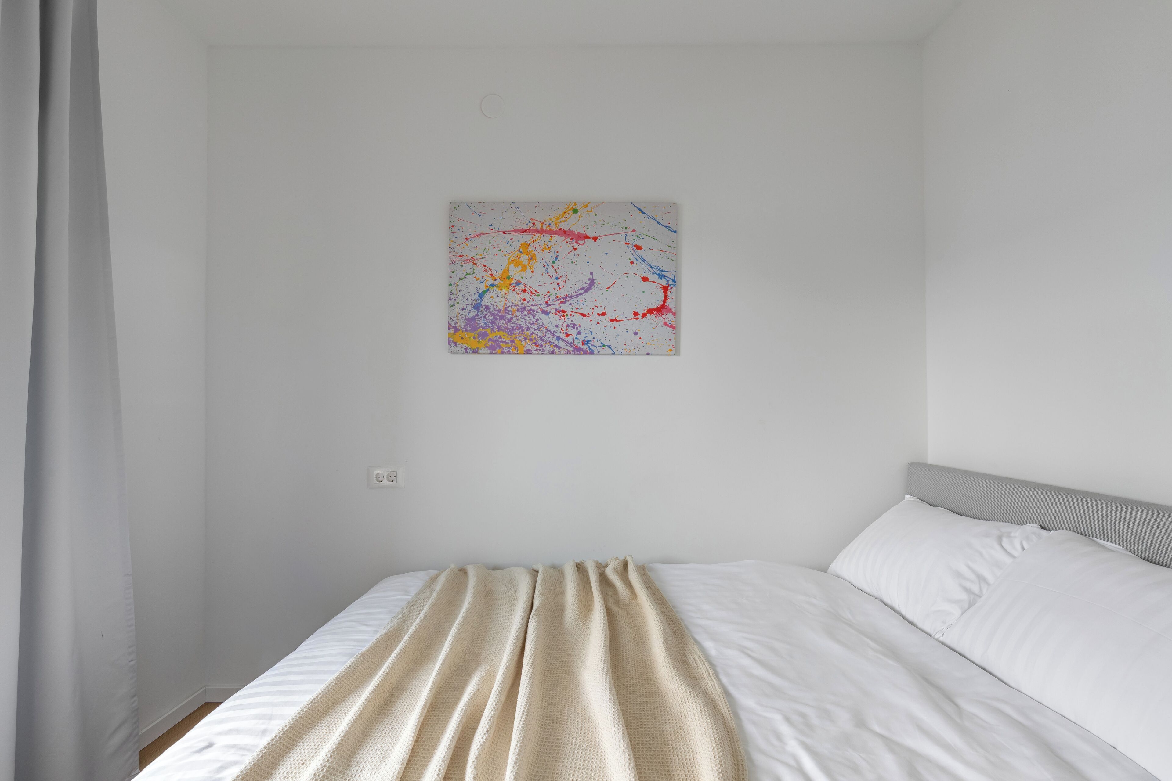 Een slaapkamer met een bed, een witte muur en een kleurrijk abstract schilderij.