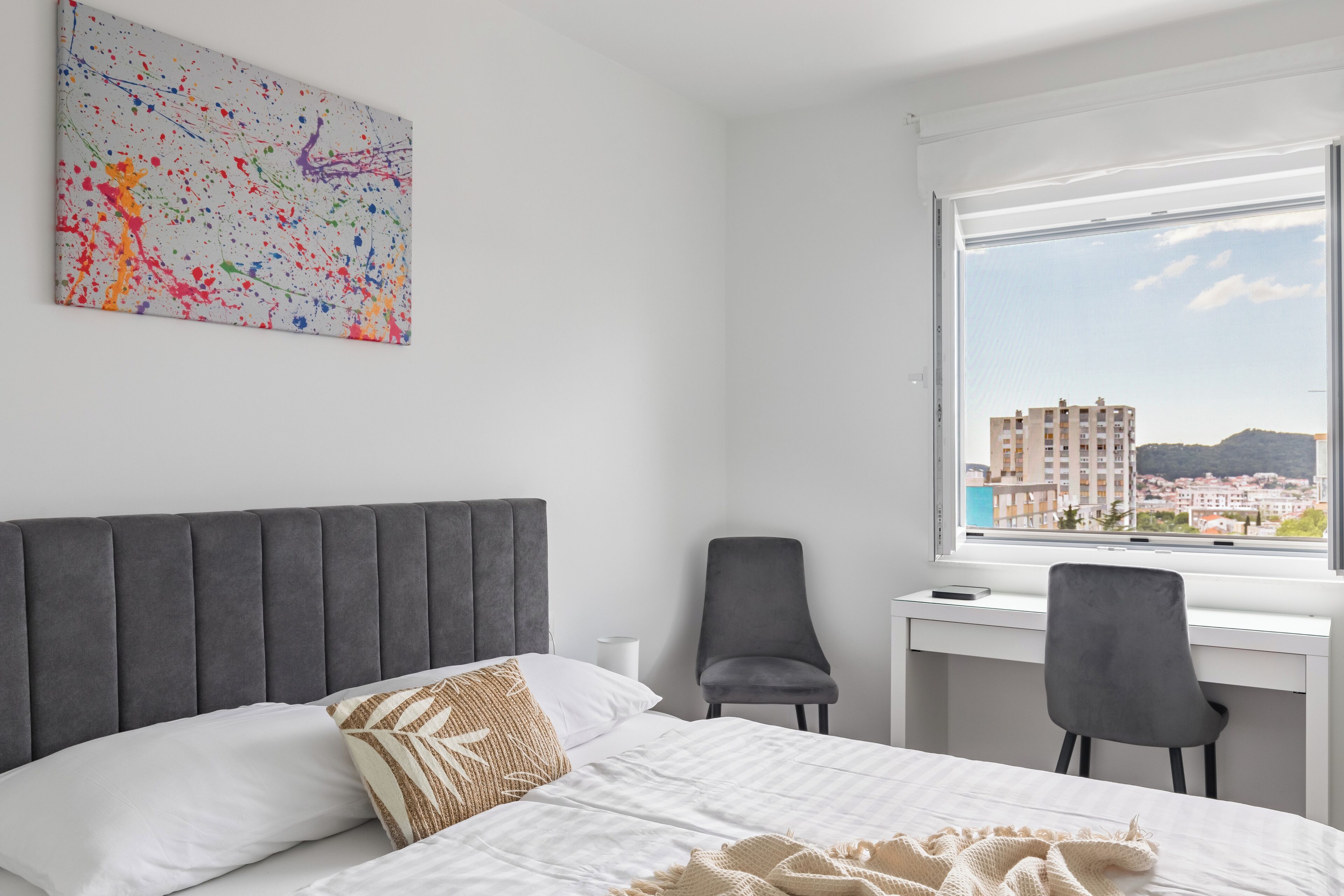 Et hotelværelse med seng, skrivebord, stol og et stort vindue med udsigt over byen.