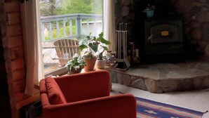 A cozy cabin interior with a stone fireplace, a wooden ceiling, and a large window overlooking a deck. - Good Vibes Cabin - Car rental available! (Craig)