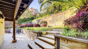 Ein überdachter Patio-Bereich mit Holzboden, Treppen zu einer Sitzgruppe und ein Garten mit verschiedenen Pflanzen und Sträuchern.