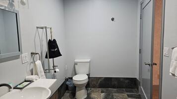 Un baño con un lavamanos blanco, un inodoro y un espejo. Hay una bolsa negra colgada en un perchero y una toalla en la pared.