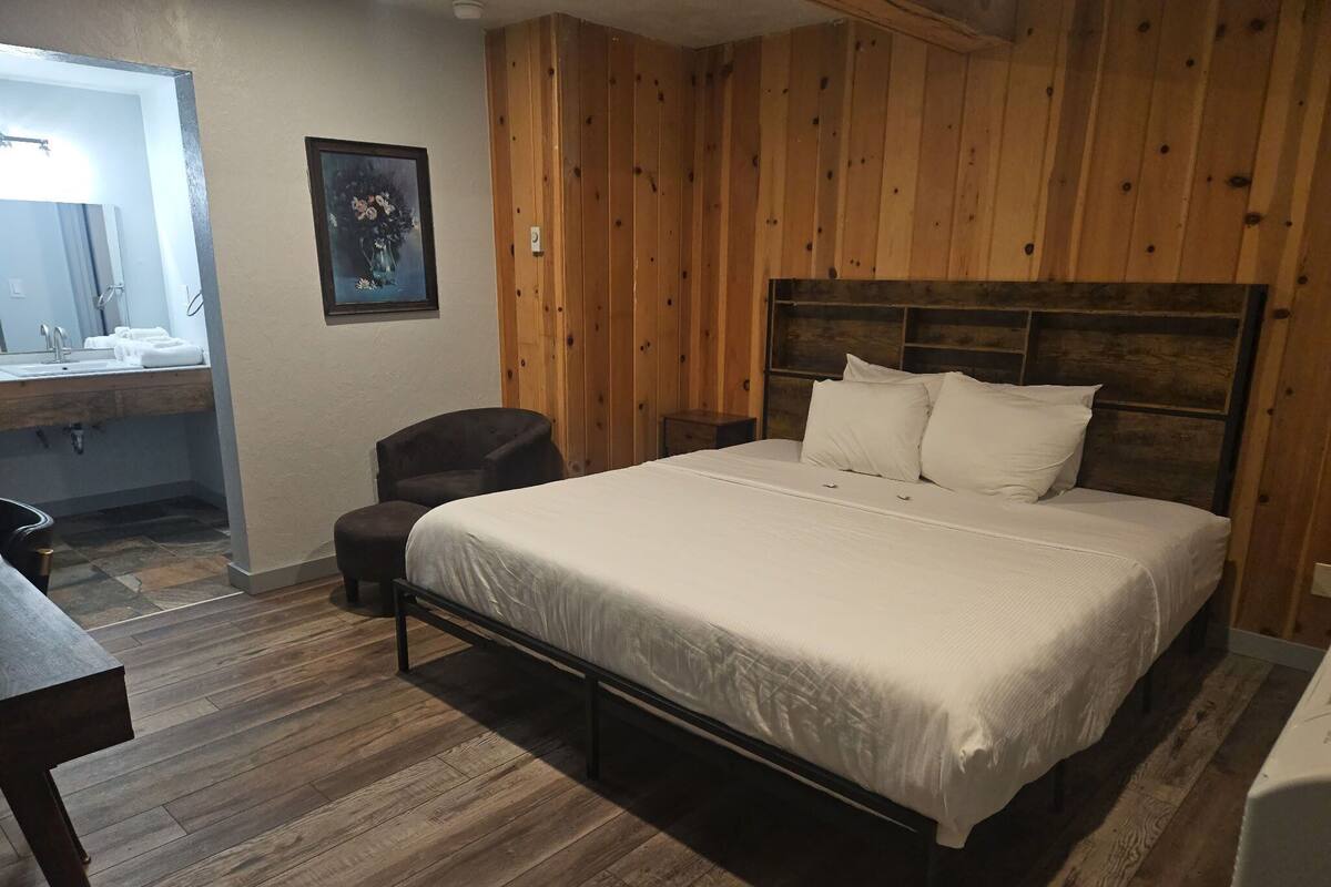 A bedroom with a wooden headboard, a large bed with white linens, a bedside table, a chair, and a wooden floor.