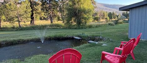 A serene outdoor setting with a small stream, red Adirondack chairs, and a wooden shed.