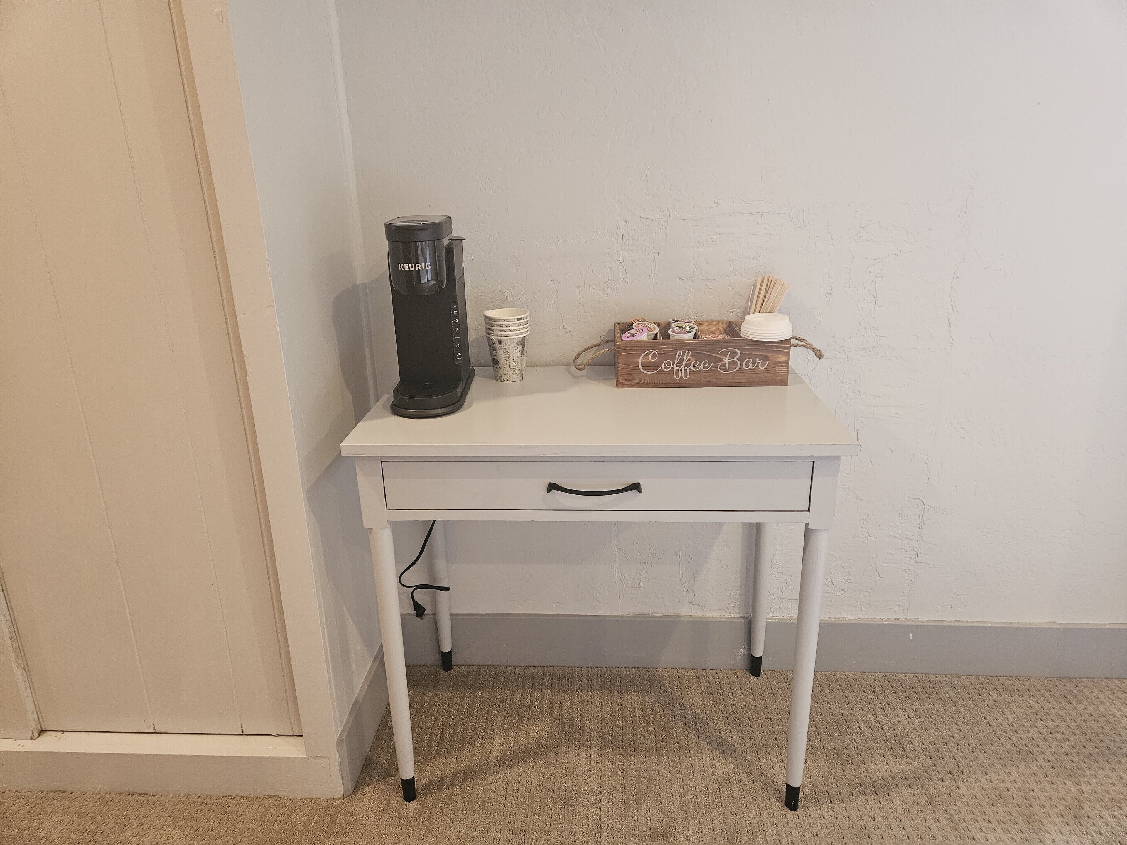 Une table blanche sur laquelle se trouvent une machine à café Keurig, une boîte contenant des accessoires pour préparer le café et une pile de gobelets en papier.