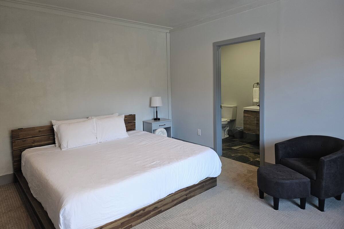 A bedroom with a bed, a nightstand, a chair, and a bathroom visible through an open door.