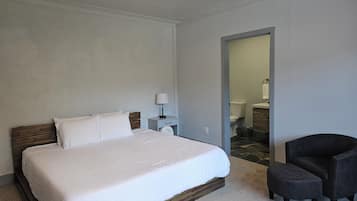 Un dormitorio con cama, mesita de noche, una silla y un baño visible a través de una puerta abierta.