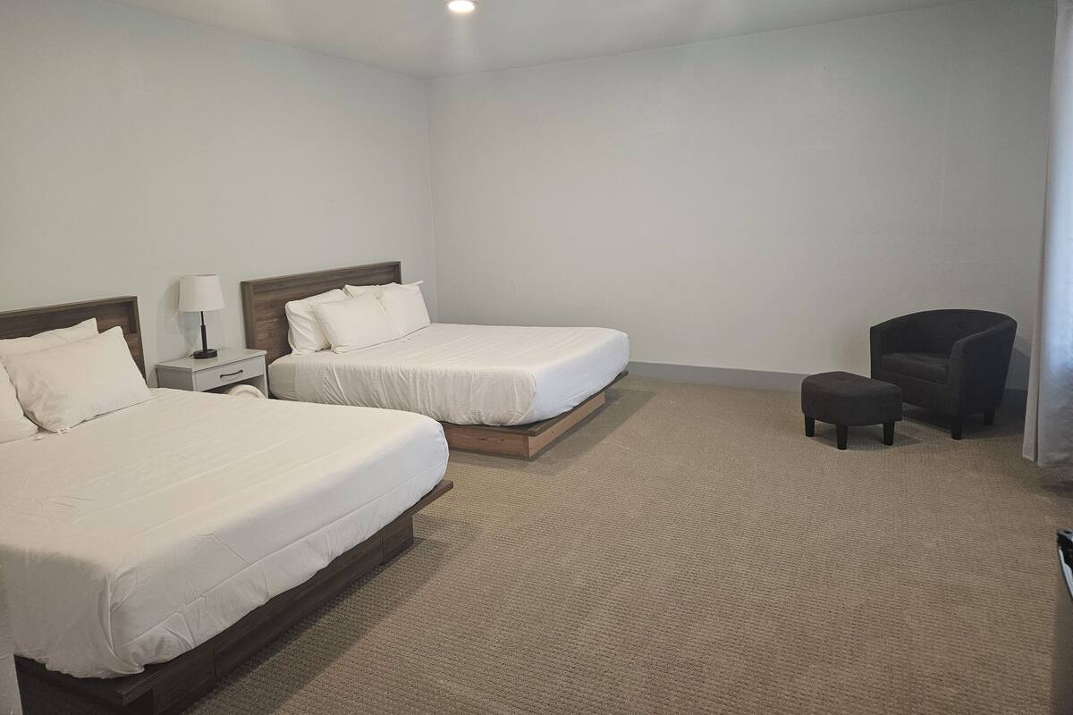 A hotel room with two beds, a nightstand with a lamp, a chair, and a carpeted floor.