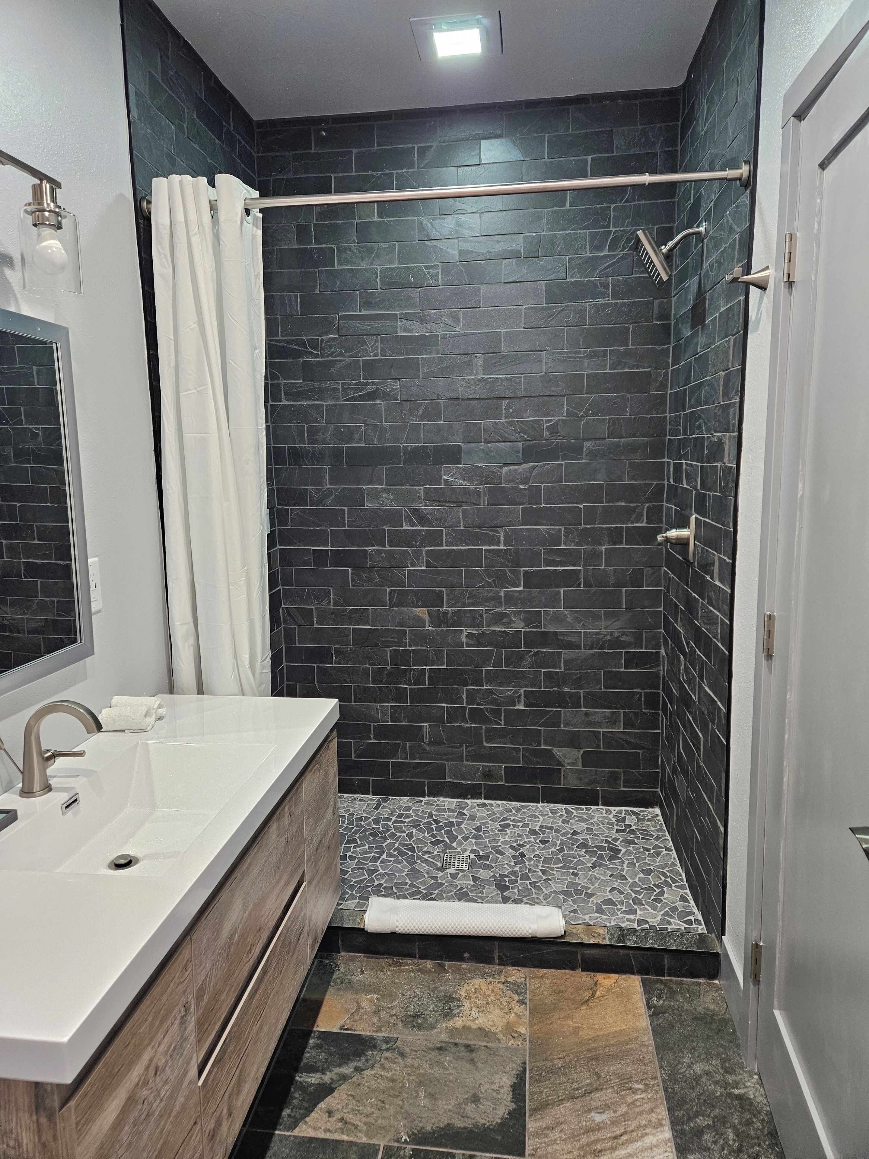 Un baño moderno con un lavamanos blanco, ducha con azulejos oscuros y tocador de madera.