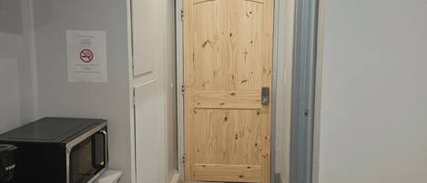 Um corredor estreito com uma porta de madeira no final, um micro-ondas numa bancada e uma lixeira.