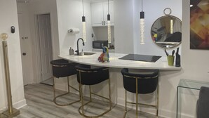 Una cocina moderna con encimera blanca, taburetes negros, un espejo grande, luces colgantes y un microondas.