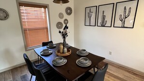 A dining area with a wooden table, black chairs, and a vase with dried flowers. There are framed pictures of cacti on the wall.