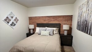 A bedroom with a bed, two bedside tables with lamps, a brick-patterned headboard, a ceiling light, and a wall-mounted decorative piece.