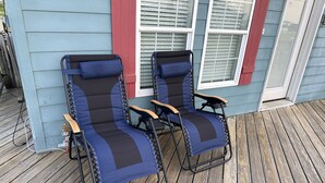 Twee terrasstoelen met metalen frame en beklede zittingen, geplaatst op een houten terras voor een huis met blauwe bekleding en rode accenten.