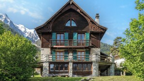 A two-story wooden house with a balcony, stone foundation, and a mountain backdrop. - Grand Chalet des Moussoux 6BR: Terrace, Garden, Views, Sleeps 10, Chamonix, France (Chamonix)