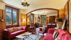 A cozy room with a red sofa, armchairs, a dining table, and a view of mountains through large windows. - Grand Chalet des Moussoux 6BR: Terrace, Garden, Views, Sleeps 10, Chamonix, France (Chamonix)