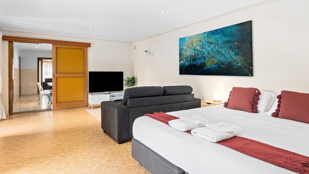A modern hotel room with a large bed, a sofa, a flat-screen TV, and a painting on the wall. - Swainson on Margaret (North Brighton)