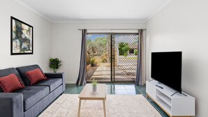 A modern living room with a grey sofa, a coffee table, a TV on a white stand, and a sliding glass door leading to a patio. - Swainson on Margaret (North Brighton)