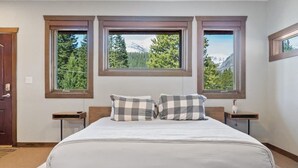 A bedroom with a bed, two nightstands, and three windows providing a mountain view.