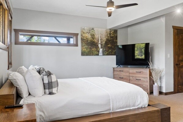 A bedroom with a bed, a TV, a ceiling fan, a window, and a painting on the wall.