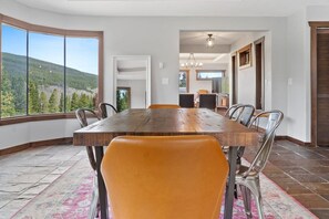 A dining area with a large wooden table, metal chairs, and a view of a forest through large windows.