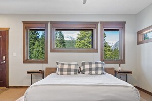 A bedroom with a bed, two nightstands, and three windows with a mountain view.