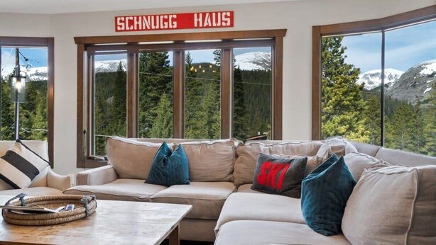 A modern living room with a large window offering a mountain view, a sofa with multiple cushions, and a wooden coffee table.