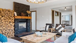 A modern living room with a stone fireplace, a stack of firewood, a chandelier, and a dining area in the background.