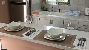 Una cocina moderna con armarios blancos, un refrigerador de acero inoxidable, una ventana con persianas y un comedor con una mesa dispuesta para dos.