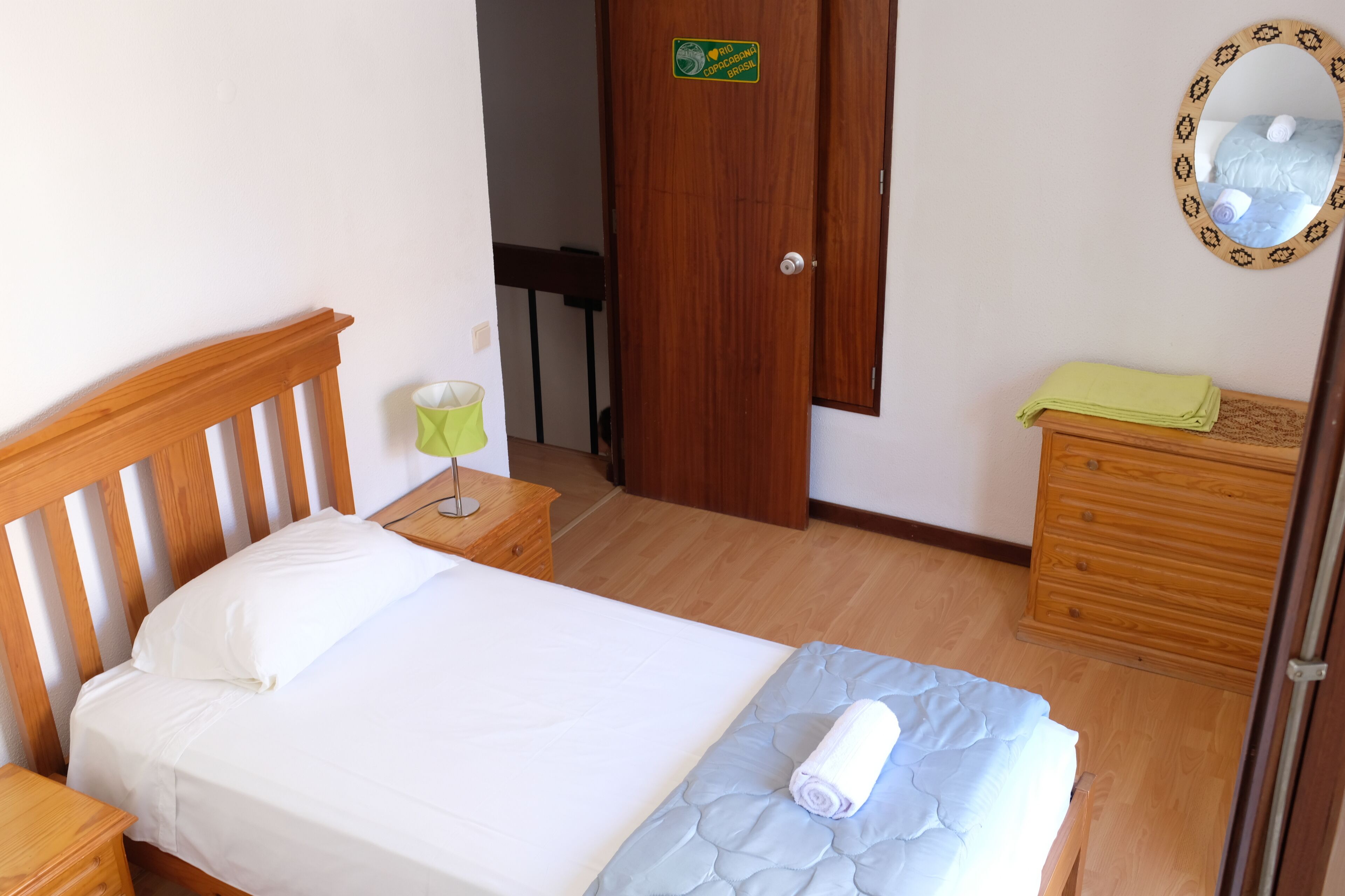 A neatly made bed with a white sheet and a blue quilt, a wooden nightstand with a lamp, a wooden dresser with a green towel, and a mirror with a decorative frame.