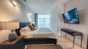 A modern hotel room with a bed, a TV mounted on the wall, a bedside table with a lamp, and a window with curtains. - Private Villa in Gated Community  (Cabo San Lucas)