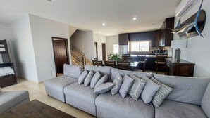 A modern living room with a grey sofa, a staircase, a kitchen with dark cabinets, and a dining area. - Private Villa in Gated Community  (Cabo San Lucas)