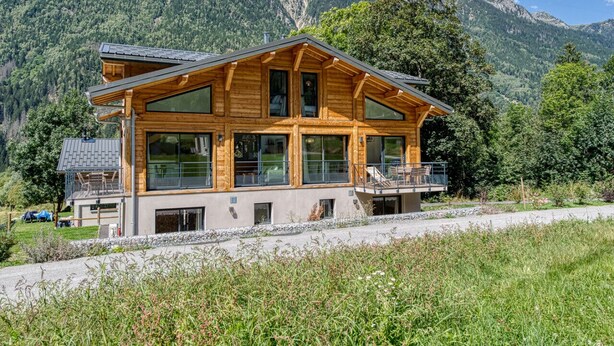 A wooden chalet with large windows, a balcony, and a mountain backdrop.