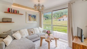 A modern living room with a grey sectional sofa, a wooden coffee table, a bookshelf, a framed picture, and a large window with a view of greenery and buildings.