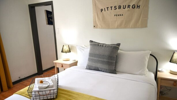 Habitación de hotel con cama, dos mesitas de noche con lámparas, un perchero para toallas y un cartel en la pared.