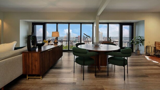 Una sala de estar moderna con un escritorio de madera, una mesa redonda con sillas, un sofá y ventanas amplias con vistas a la ciudad.