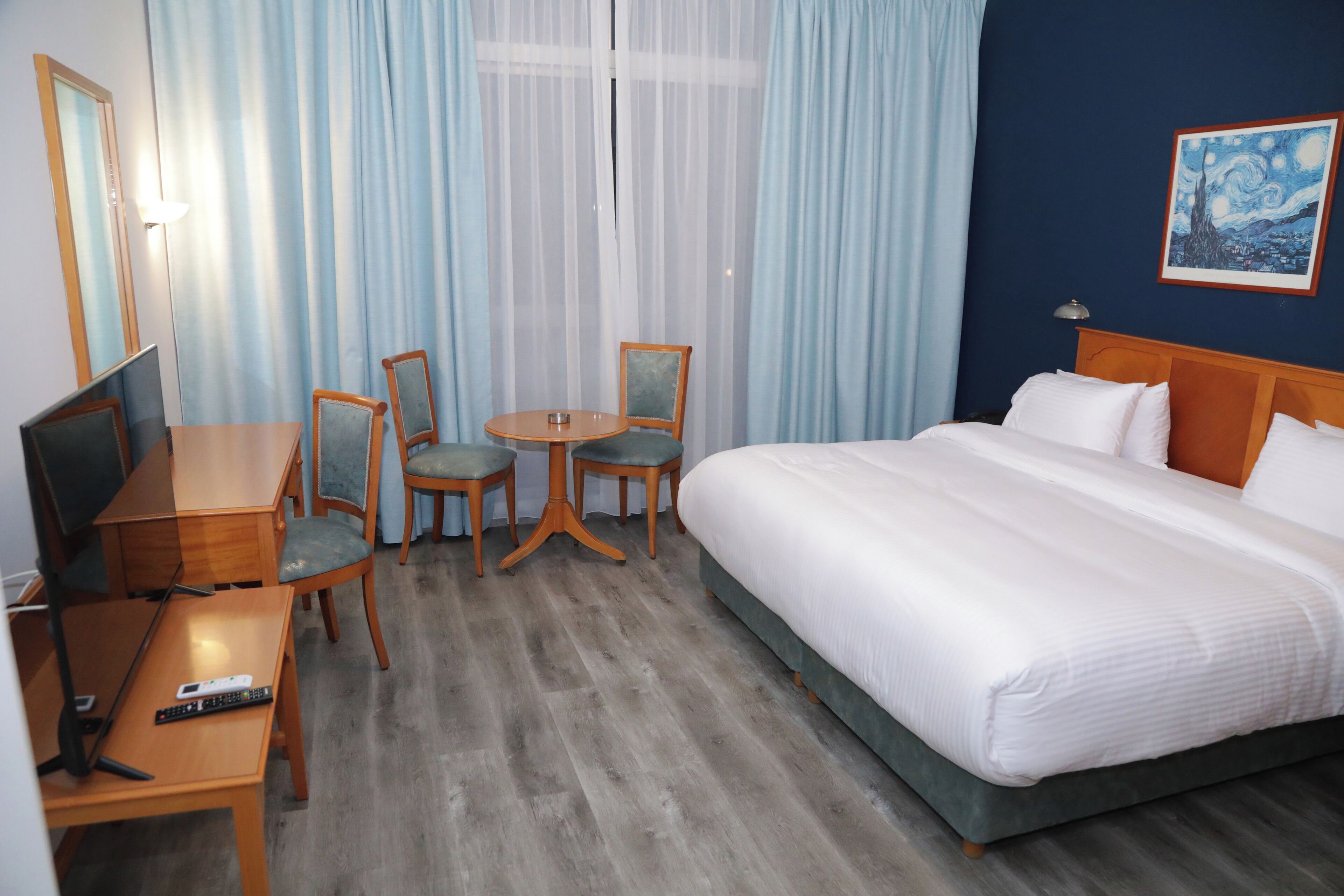 A hotel room with a bed, a desk, two chairs, a TV, and a painting on the wall.
