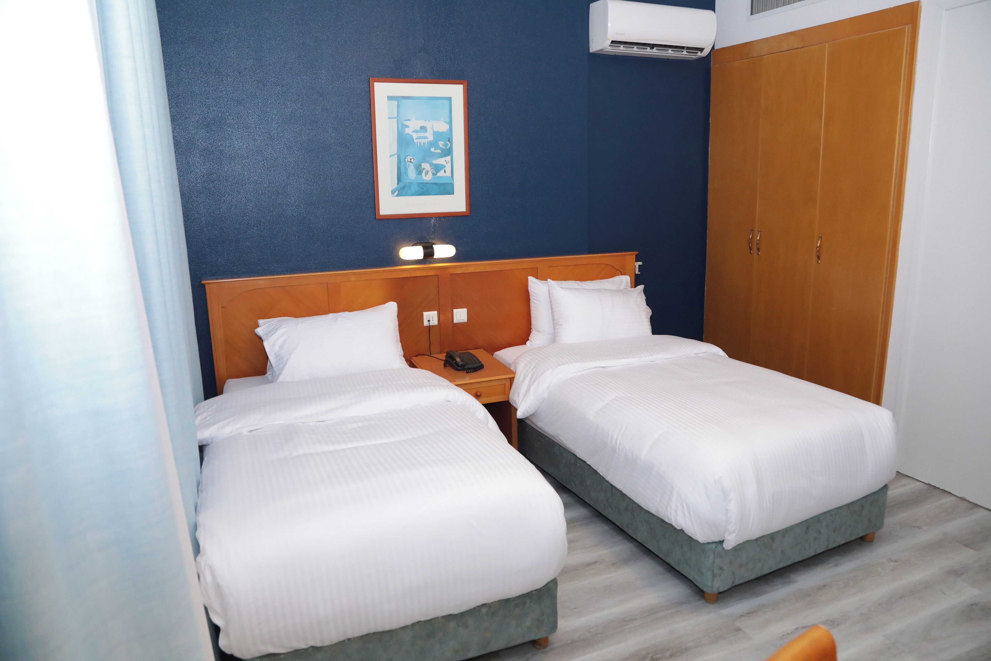 Een hotelkamer met twee enkele bedden, een houten hoofdeinde, een nachtkastje met een telefoon, een lijst aan de muur en een kledingkast.