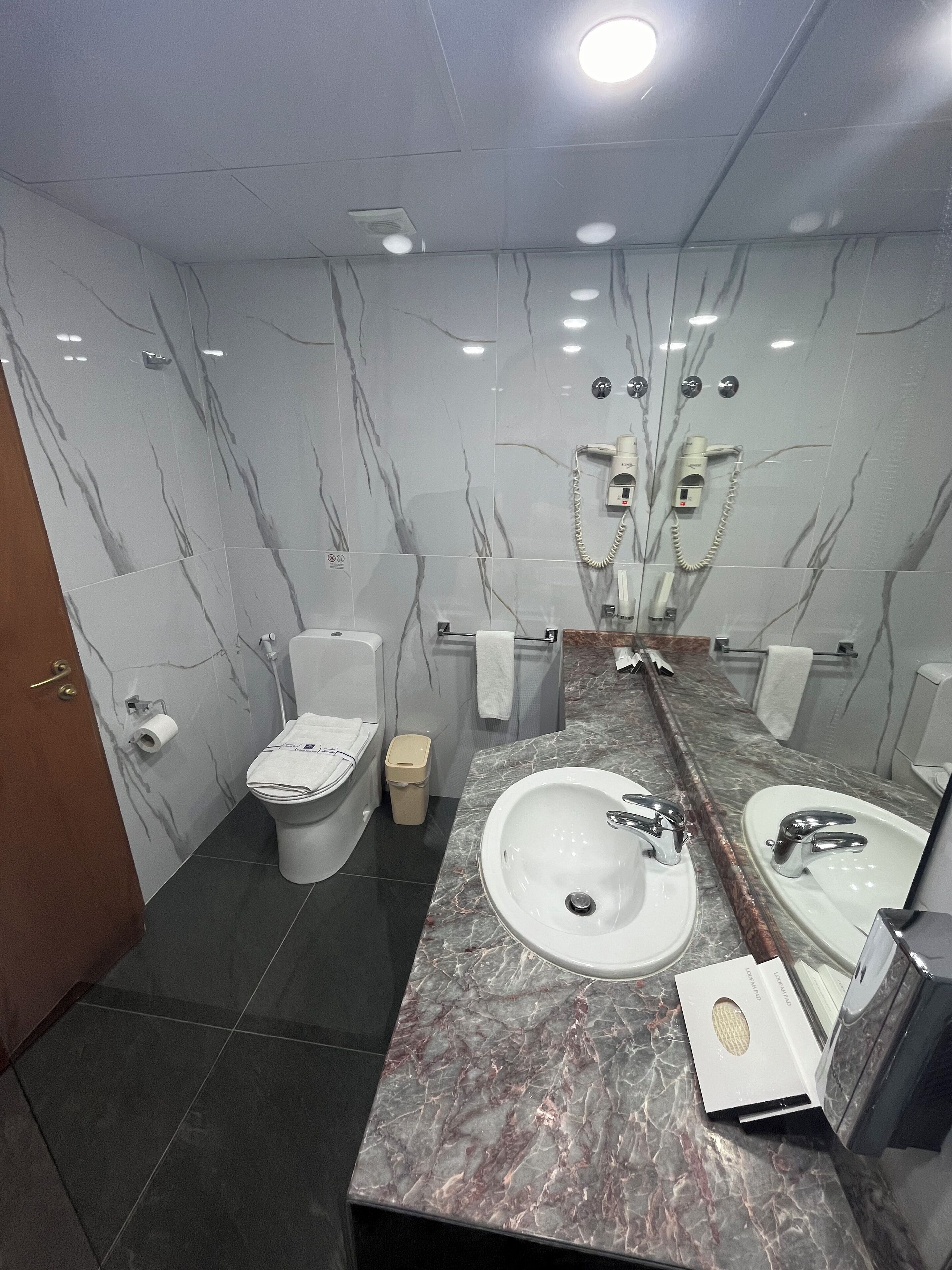 A modern bathroom with marble walls, a large sink, two toilets, and a trash bin.