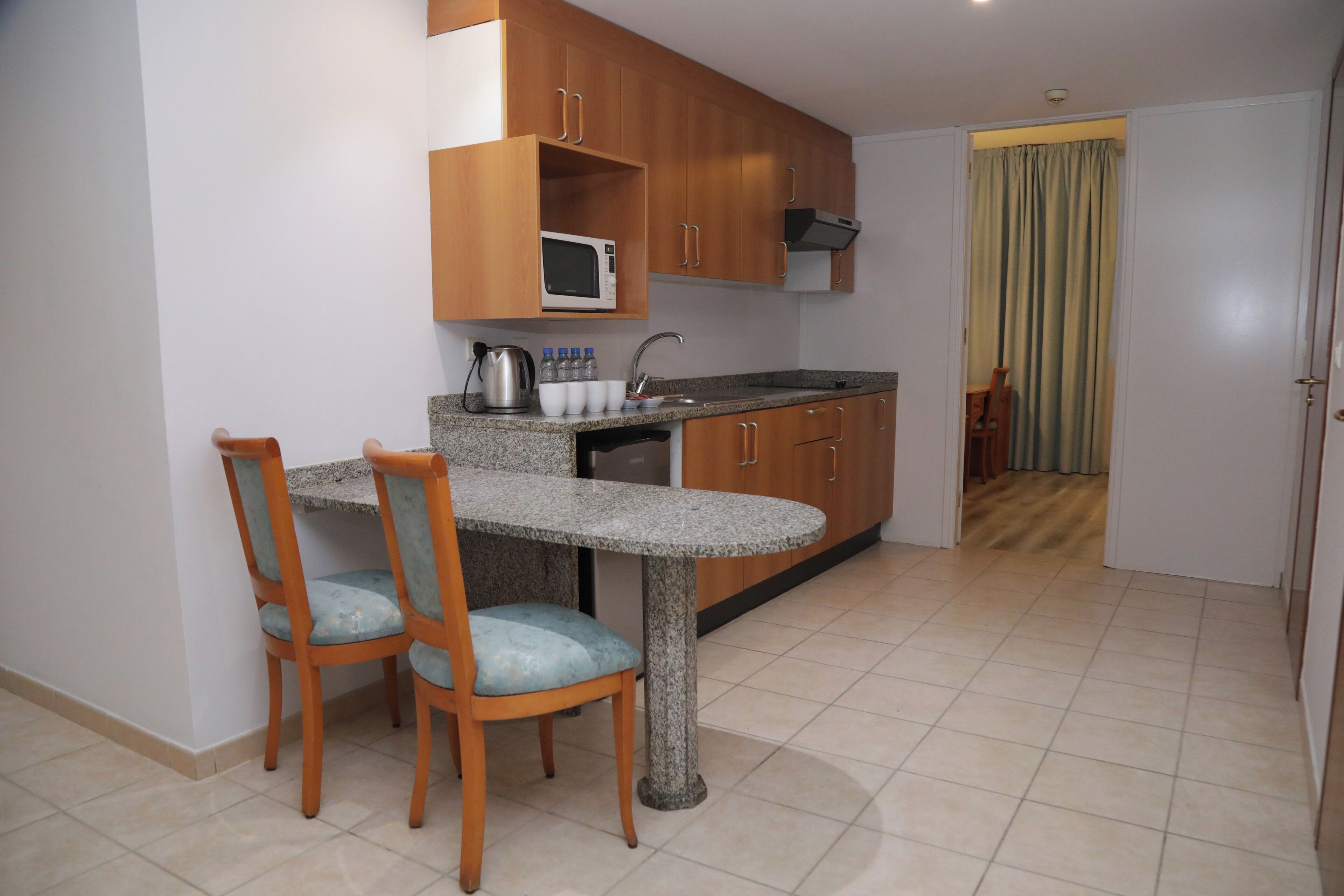 A compact kitchen with wooden cabinets, a granite countertop, a microwave, and a sink. There are two chairs with blue cushions.