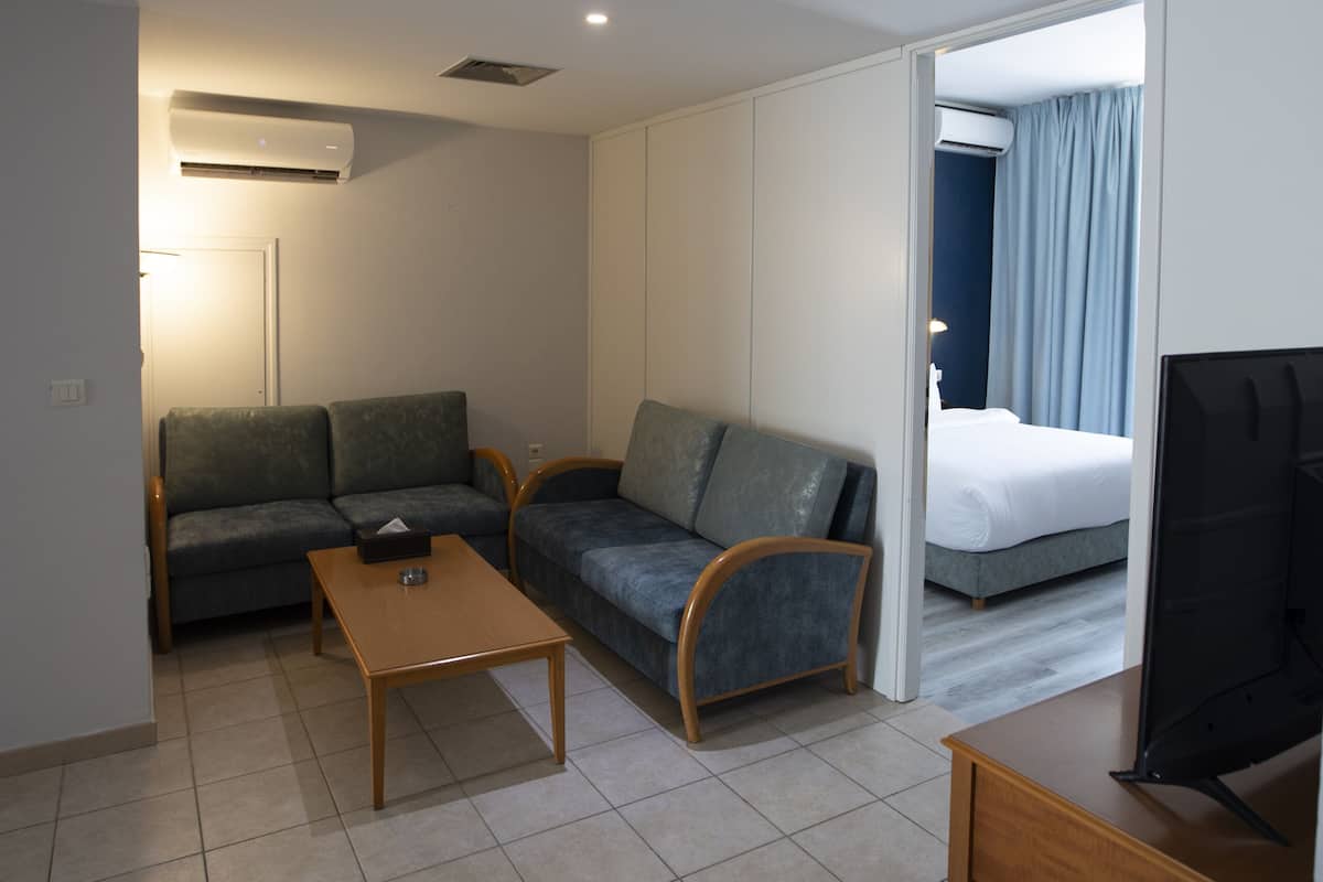 A modern hotel room with a sofa, a small table, a TV, and a separate bedroom area with a bed and curtains.