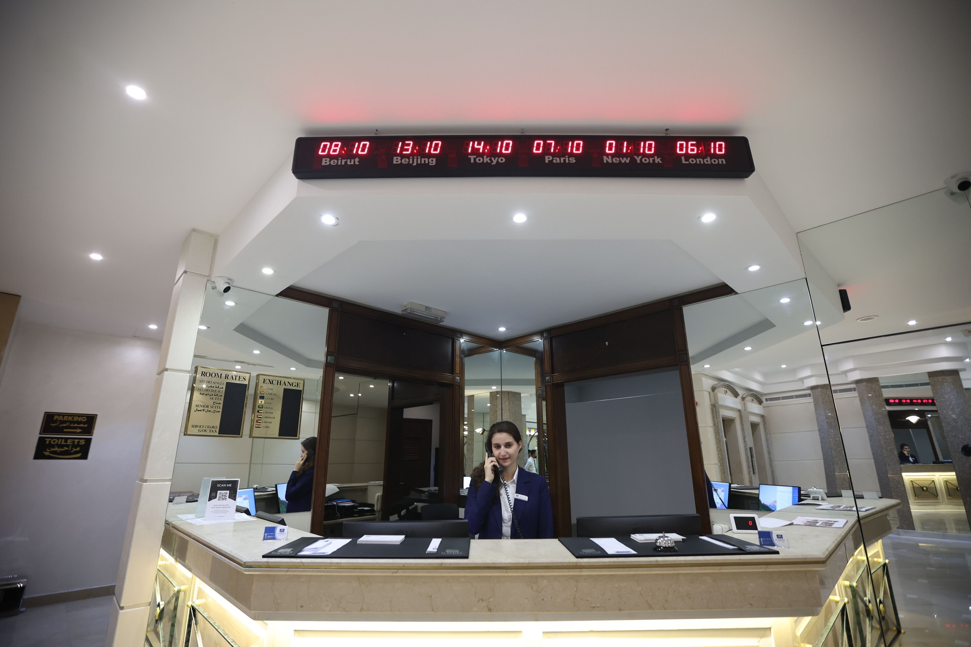 A hotel reception desk with a digital clock displaying time in multiple cities, a glass partition, and a receptionist.