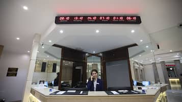 A hotel reception desk with a digital clock displaying time in multiple cities, a glass partition, and a receptionist.