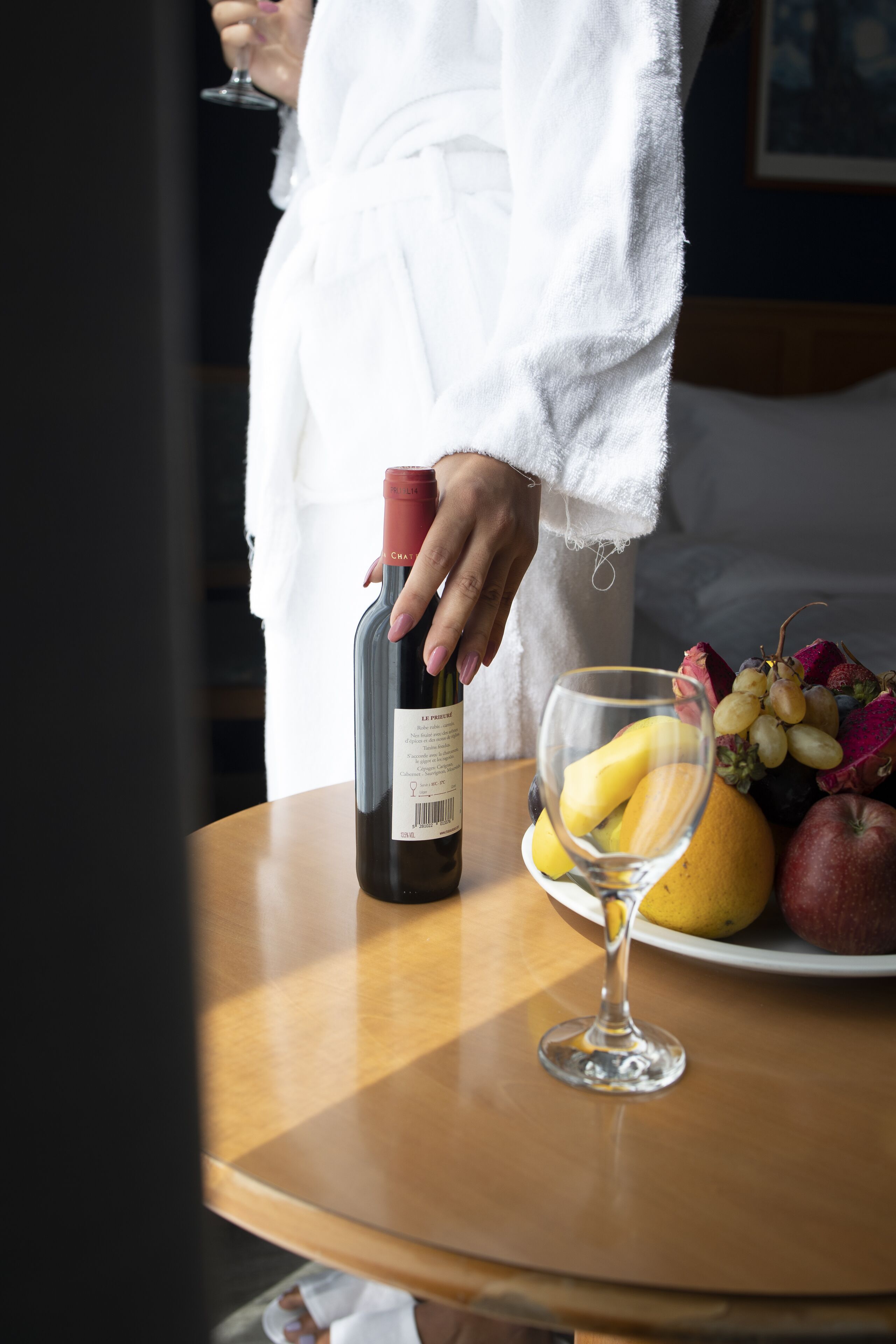 Een persoon in een witte badjas die een fles wijn vasthoudt, staand naast een tafel met een wijnglas en een schaal fruit.