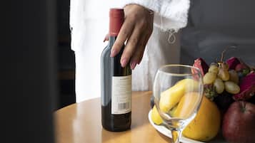 Een persoon in een witte badjas die een fles wijn vasthoudt, staand naast een tafel met een wijnglas en een schaal fruit.