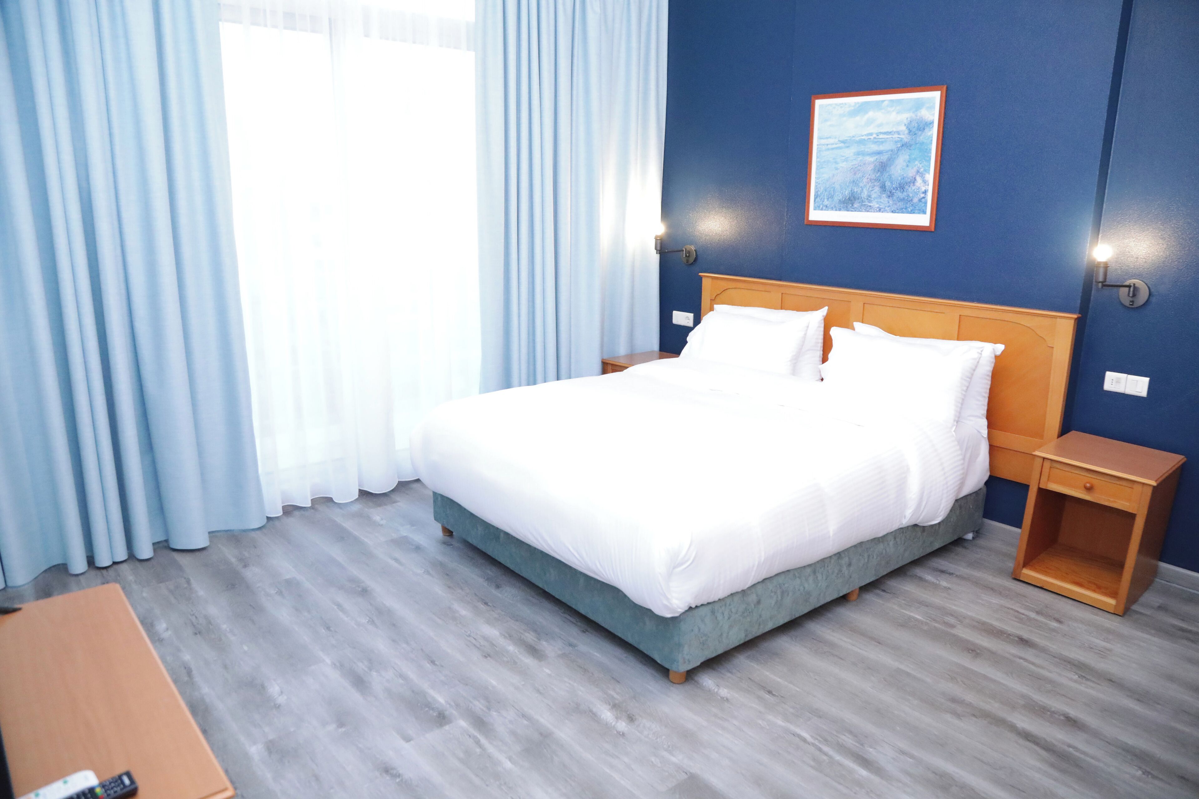 A hotel room with a bed, a nightstand, a wooden table, and a painting on the wall.