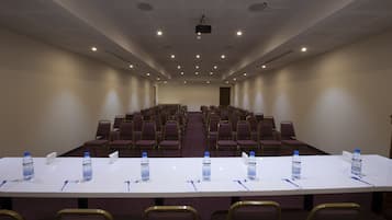 Un salón de conferencias con filas de sillas, una larga mesa con botellas de agua y bolígrafos, y un proyector en el techo.