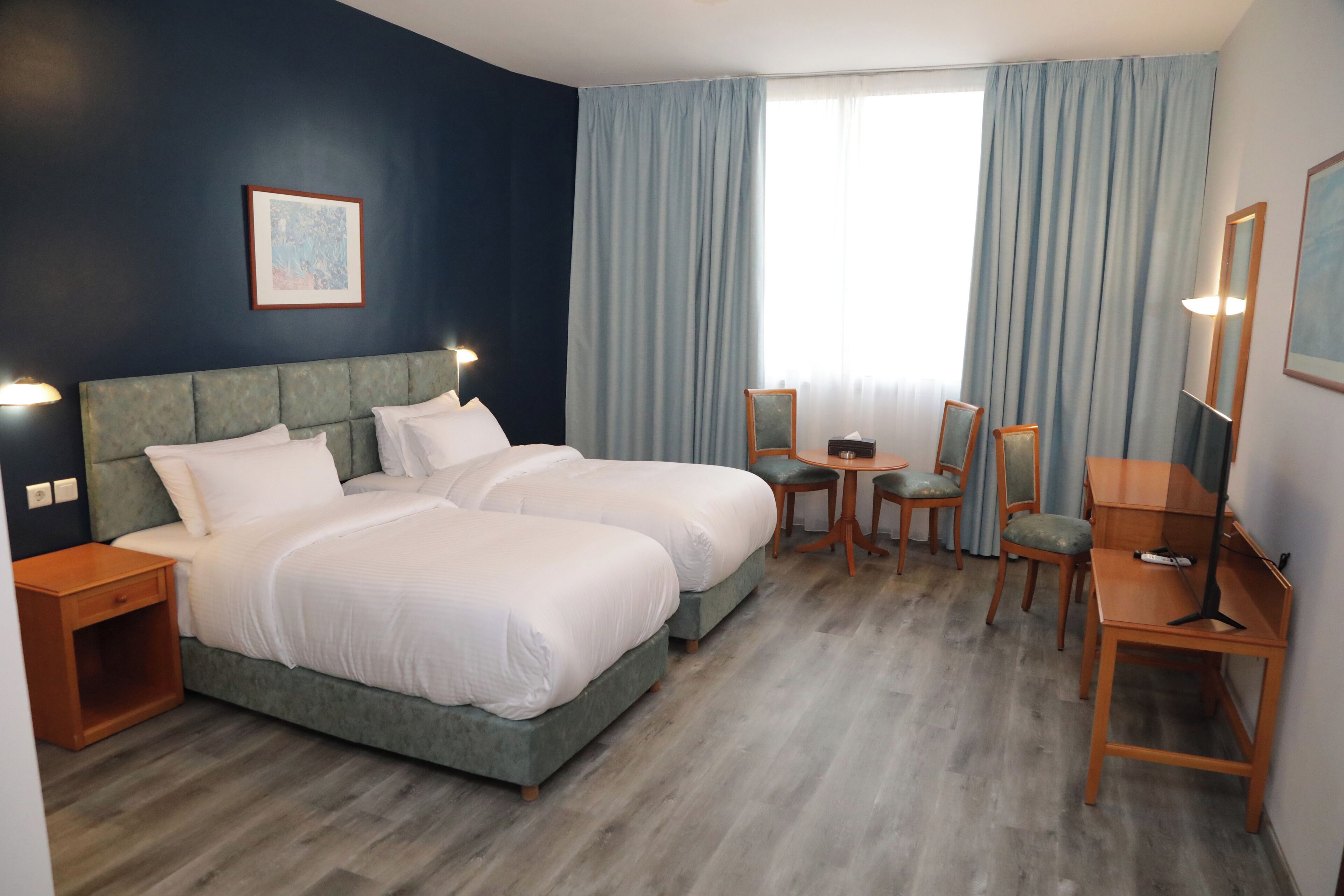 Ein Hotelzimmer mit zwei Betten, einem Schreibtisch, Stühlen, einem kleinen Tisch und einem großen Fenster mit Vorhängen.