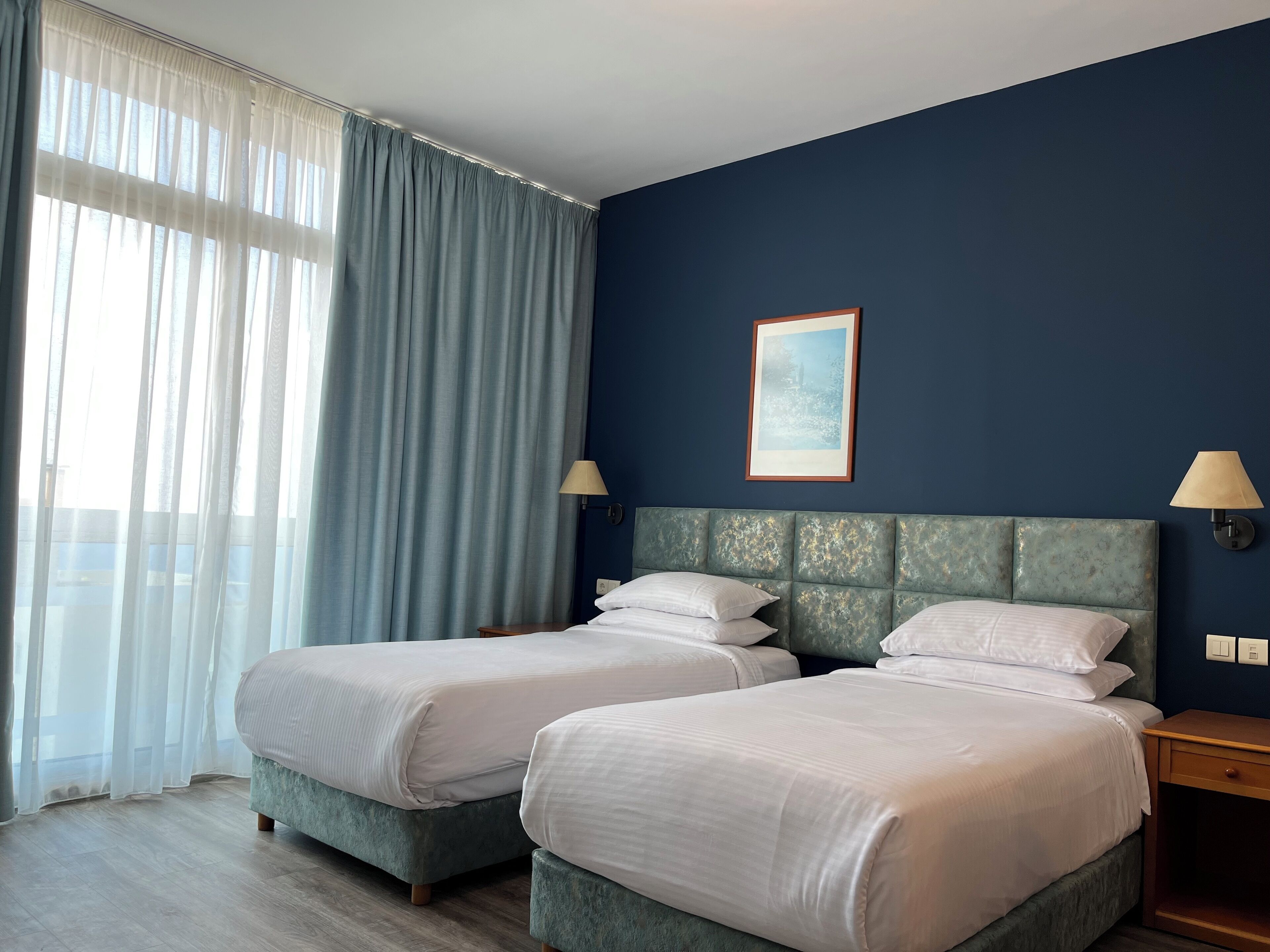 A hotel room with two beds, a large window with sheer curtains, a framed picture on the wall, and a nightstand with a lamp.