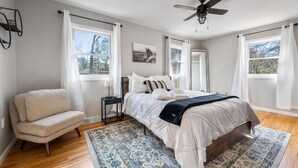Un dormitorio con una cama, una silla, un ventilador de techo y una ventana con cortinas.